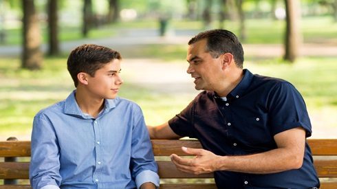Father talking to teen son while sitting on a bench in a park